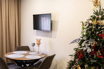 cozy living room features a round wooden table with lace doilies, a wall-mounted TV, and a beautifully decorated Christmas tree with red and gold ornaments, creating a festive and warm atmosphere