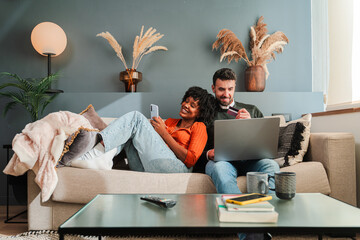 A couple happily engaged in online shopping, using a laptop and smartphone to browse products while...