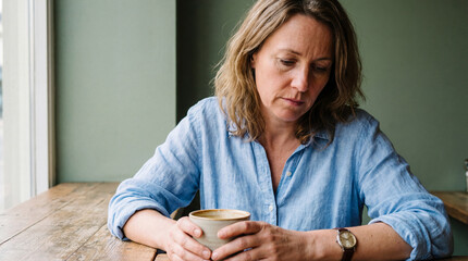 Middle aged woman sitting alone at wooden table holding coffee cup, looking down with thoughtful and lonely expression indoors