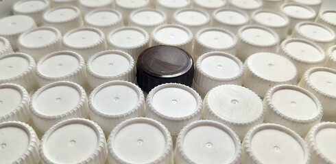 A field of uniform white plastic bottle caps, tightly packed, with a single dark brown/black cap standing out in the center, creating a contrast pattern
