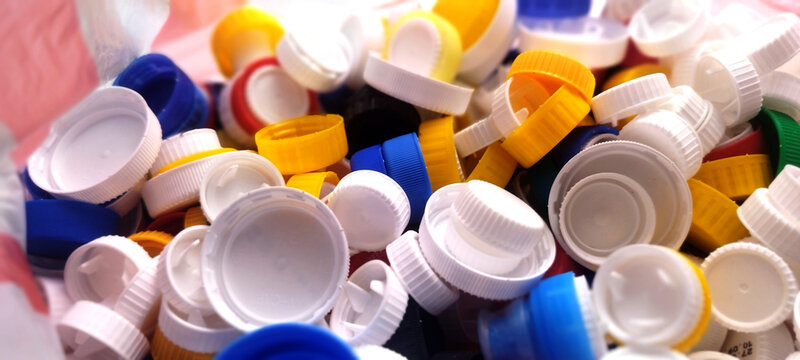 A close-up, high-angle shot of a large pile of assorted plastic bottle caps, predominantly white, with splashes of blue, yellow, and red, suggesting recycling or sorting