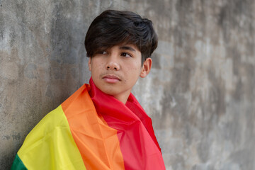 Teenaged boy covers his body with rainbow flag, lgbtqai symbol, standing and looking ahead in front of wall, concept for calling out people to respect human rights of lgbtqai people around the world.