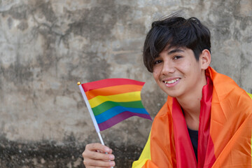 Teenaged boy has rainbow flag on body and showing rainbow flag, lgbtqai symbol, in front of wall, concept for calling out all people to respect human rights of lgbtqai people around the world.