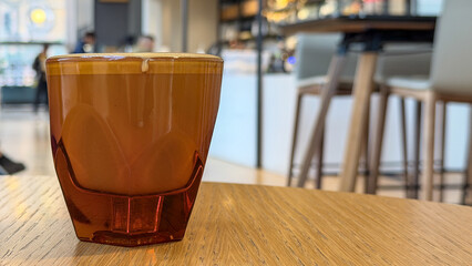 Flat white coffee beverage in transparent orange-tinted, faceted glass cup on wooden café table with blurred interior in background