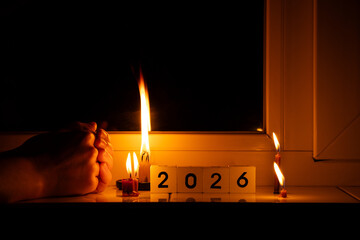 Cubes with the number 2026, burning candles, and women's hands on a windowsill in a dark apartment, symbolizing life without electricity, blackouts, and power outages in Ukraine during