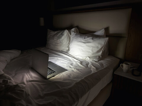 Laptop on hotel bed at night with soft light, empty bedroom interior showing late work, remote lifestyle or insomnia concept, quiet modern hotel room with technology and solitude