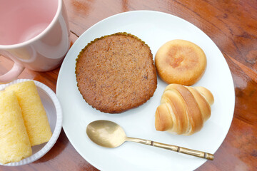 Assorted baked goods on a white plate including cake, Bakery set, Round Bun, and mini croissant with a gold spoon and pink cup, ideal for food photography and menu use, Top view
