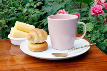 Breakfast setup with a pink coffee cup, pastries, and snacks on a wooden table in a rose garden. Warm, natural outdoor scene ideal for food and lifestyle themes