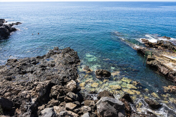 Fototapeta premium Rocky volcanic shoreline with clear turquoise Atlantic water and swimmers near natural bathing area, Tenerife, Canary Islands, Spain.