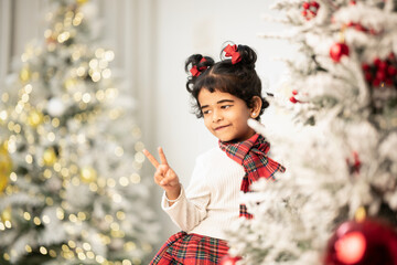 Cute young girl posing playfully near a Christmas tree, wearing red and white holiday clothing, enjoying magical winter celebration indoors