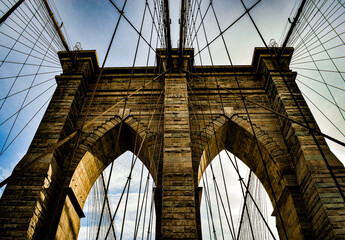Dramatic architectural view of the Brooklyn Bridge, captured from below with strong lines, stone arches and converging cables creating a powerful geometric composition.
