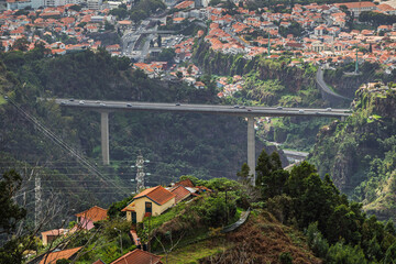 City view of Funchal in Madeira. Arial view of a long concrete bridge crossing a deep green valley in Madeira, with dense city neighborhoods and red-tiled roofs in the background
