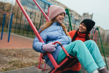 A happy, smiling teenage girl swings on a swing at a playground outdoors with her caring young mother. Childhood concept photo.
