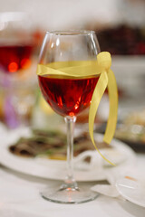 Glass of red wine decorated with yellow ribbon standing on festive table with blurred background. Elegant celebration detail, restaurant setting, romance, anniversary, holiday concepts.
