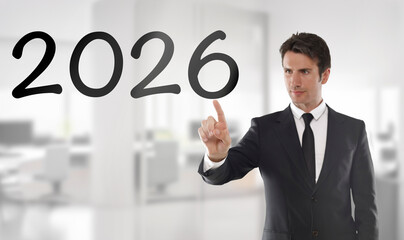 Professional businessman in a dark suit points toward large 2026 numbers displayed on a transparent virtual screen, set against a bright blurred modern office interior. 