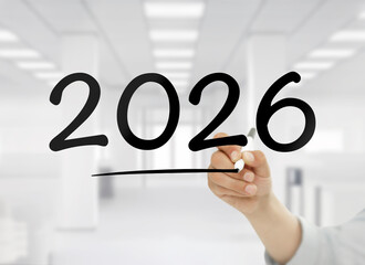 Close up of a human hand using a marker to write the year 2026 with an underline on a transparent screen, set against a bright blurred modern office interior. 