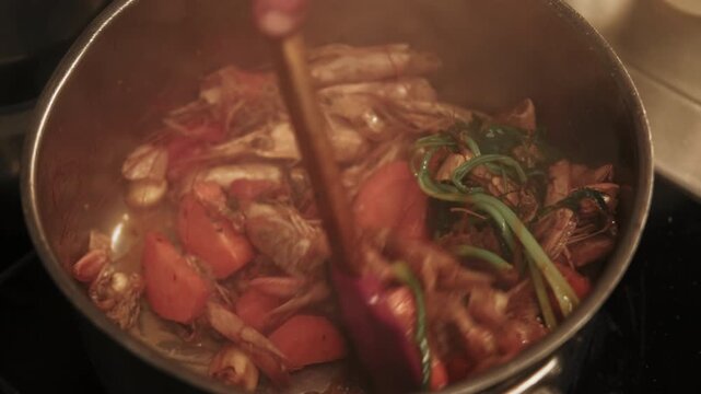 Cooking seafood. Shrimp shells. Chef hand stirring fried ingredients for bisque soup. Tom yum kung with carrots green herbs spices in hot steaming pot.