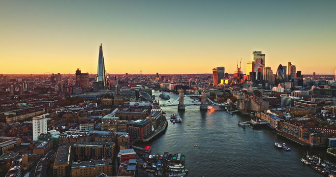 England, aerial London skyline at sunset with iconic landmarks Tower Bridge, The Shard skyscraper, and illuminated buildings of Canary Wharf on River Thames. Modern cityscape under bright evening sky