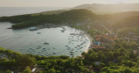 Padang Bai harbor on Bali island at sunset, fishing boats and ferries anchored in bay, coastal town nestled among lush green hills under soft light. Tropical paradise. Aerial view drone panorama © Goinyk