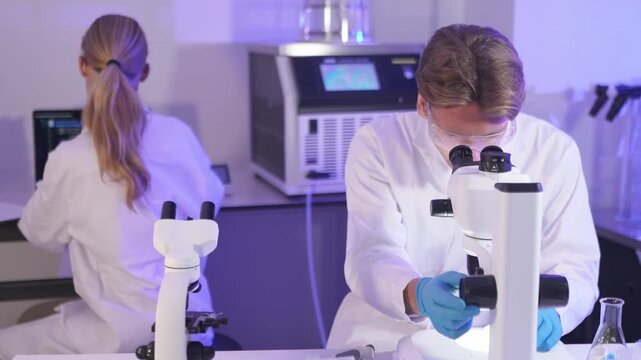 Scientist examines samples with microscope in laboratory setting while colleague works on computer