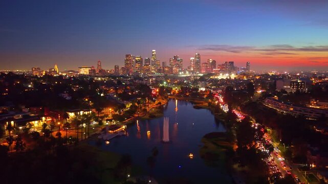 A dramatic aerial view at night captures the brilliant, illuminated skyline of downtown Los Angeles reflecting across the dark, placid surface of Echo Park Lake