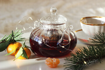 A transparent teapot sits on a small table. Nearby are tangerines, a fir branch, and a garland.