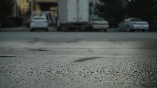 Low-angle view of damp, gritty asphalt in a quiet urban parking area, with blurred parked cars and a box truck in the background, revealing texture, potholes, and evening bokeh.