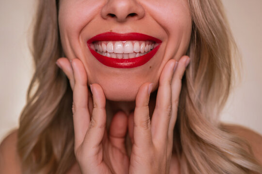 Close up of a smiling blonde woman with bright red lipstick and perfect white teeth. Hands touching face, healthy skin, beauty and dental care concept, confident feminine smile