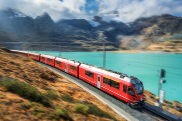 Modern red train is moving along railway in swiss alps mountain range near lake on sunny day with cloudy sky in autumn. Landscape. Bernina Express. High speed train with motion blur effect. Travel