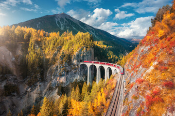 Aerial view of modern red train on Landwasser viaduct in alpine mountains, colorful forest at sunset in autumn. Bernina Express, Switzerland. Top view of train, railroad, orange and red trees in fall