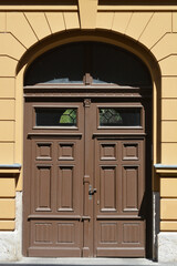 A large, symmetrical, brown wooden double door with panels and an arched transom window is set within a decorative yellow stone facade