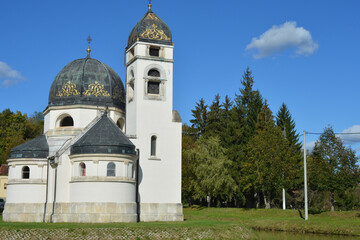 The Greek-Catholic Church of the Annunciation in Strmac Pribicki, Croatia, with its white facade and dark domes, is situated beside a pond and green trees under a blue sky