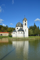 A full-view shot captures the Greek-Catholic Church of the Annunciation in Strmac Pribicki, Croatia, standing by a pond, beautifully reflected in the green water under a blue sky