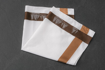 A neatly folded white cotton handkerchief with a wide brown and tan border, featuring a sailboat pattern on a dark gray, textured surface.