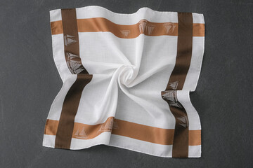 A white cotton handkerchief with a wide brown and tan border, featuring a sailboat pattern is folded into a swirl pattern and placed on a dark gray surface.