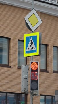 Urban pedestrian crossing traffic light with digital countdown timer switching from red stop to green go beneath priority and crosswalk signs beside brick building facade