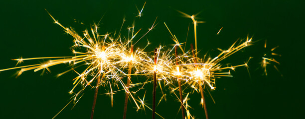 Burning sparklers illuminate a dark green background. The bright sparks create a magical holiday atmosphere: New Year's, Christmas, or a party. A background image for cards and design. Five sparklers.