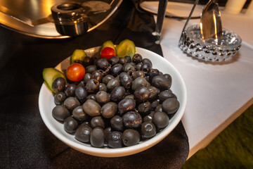 A white serving bowl of black olives garnished with cherry tomatoes and cucumber slices on a buffet table.
