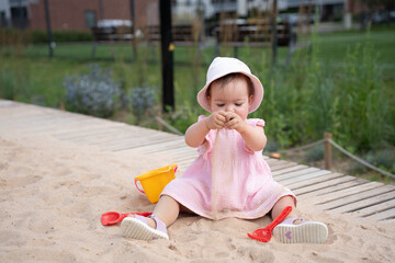 young child in pink dress and sun hat playing joyfully with sand in a green park setting, surrounded by a yellow bucket and red toy shovel on a sunny day, peaceful, colourful, bright, serene, scene