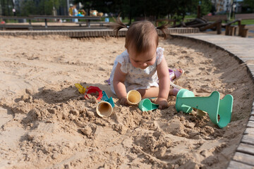 young child in white outfit plays intently in a sunny sandbox with colorful toys, surrounded by a circular wooden border, in a lively park setting on a bright day, toddler, pure, hands, urban, path