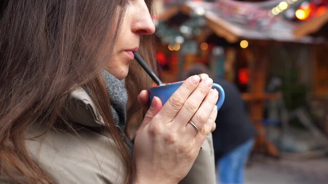 Young woman drinking hot cocoa at a Christmas market. Cozy winter atmosphere, festive lights and decorations in the background. Warm drink with whipped cream in hands, cold weather, holiday mood, slow