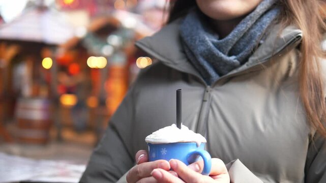 Young woman drinking hot cocoa at a Christmas market. Cozy winter atmosphere, festive lights and decorations in the background. Warm drink with whipped cream in hands, cold weather, holiday mood, slow
