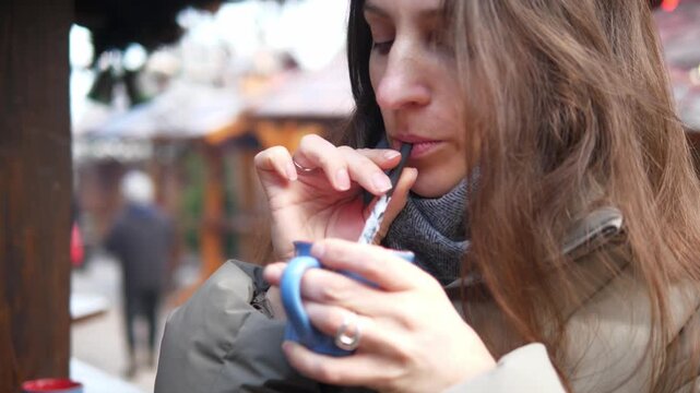 Young woman drinking hot cocoa at a Christmas market. Cozy winter atmosphere, festive lights and decorations in the background. Warm drink with whipped cream in hands, cold weather, holiday mood, slow