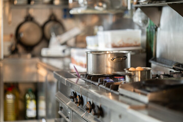 Stovetop with stainless steel saucepans on lit gas burner against blurry background of restaurant kitchen