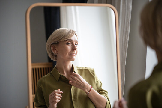 Beautiful middle aged woman getting ready for date or daily walk, standing in front of mirror and fixing clothing outlook in reflection, checking new trendy blouse with positive satisfied expression.