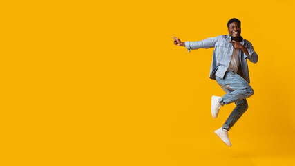 Emotional african american millennial man jumping up and pointing at copy space over yellow studio background, happy black guy recommending nice offer, showing blank space for advertisement, panorama