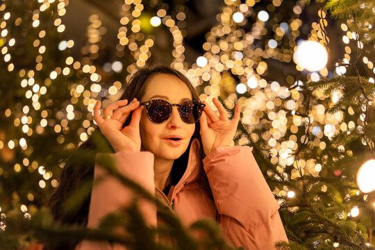 A surprised girl wearing sunglasses at a Christmas market in Europe. In the background, bokeh from the lights, with reflections of the market lights in her sunglasses.
