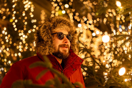 A surprised bearded man wearing a red jacket at a Christmas market in Europe. In the background, bokeh from the lights, with reflections of the market lights in his sunglasses.
