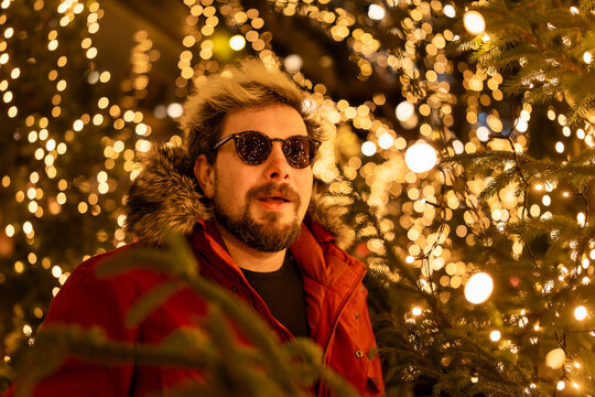A surprised bearded man wearing a red jacket at a Christmas market in Europe. In the background, bokeh from the lights, with reflections of the market lights in his sunglasses.