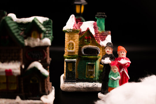 Cozy miniature Christmas village scene with caroling figures singing beside a snow-covered house and trees. Warm glowing windows, soft focus, and dark background create a nostalgic holiday atmosphere.